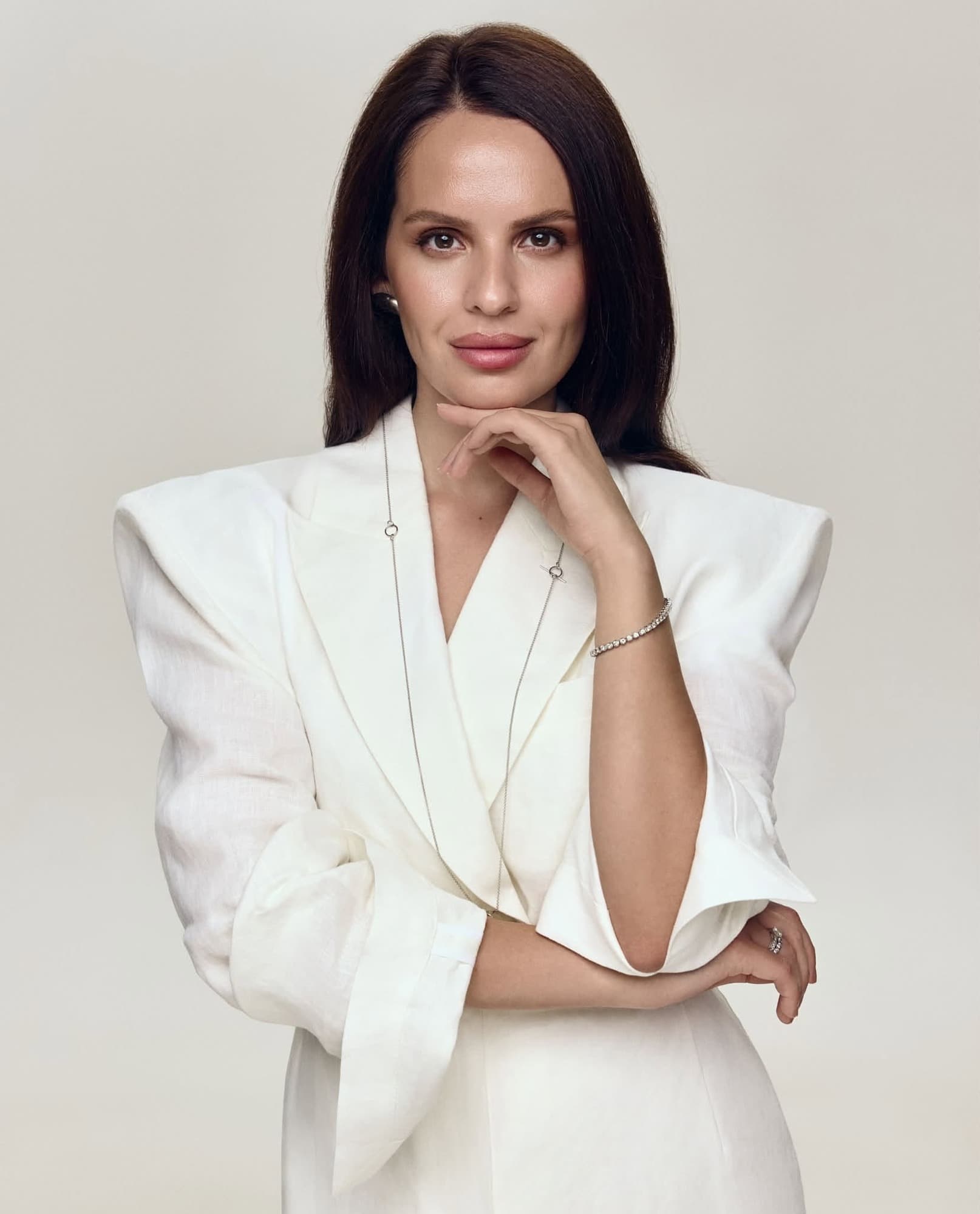 Anna Bon in a white structured jacket against a pale backdrop, hand to chin.