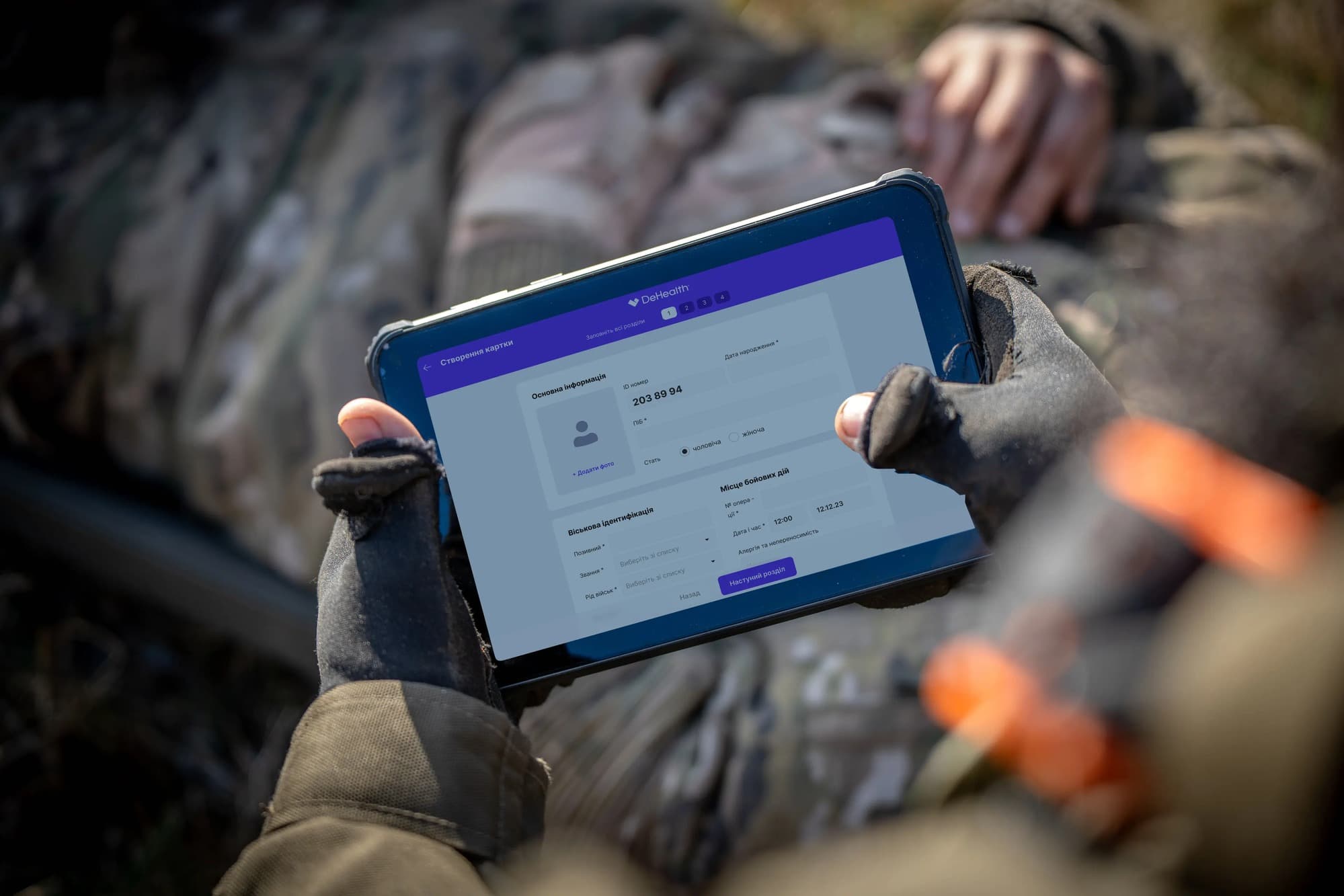 A medic in gloves holding a rugged tablet showing the Army Health System record form, with a casualty lying beside them.