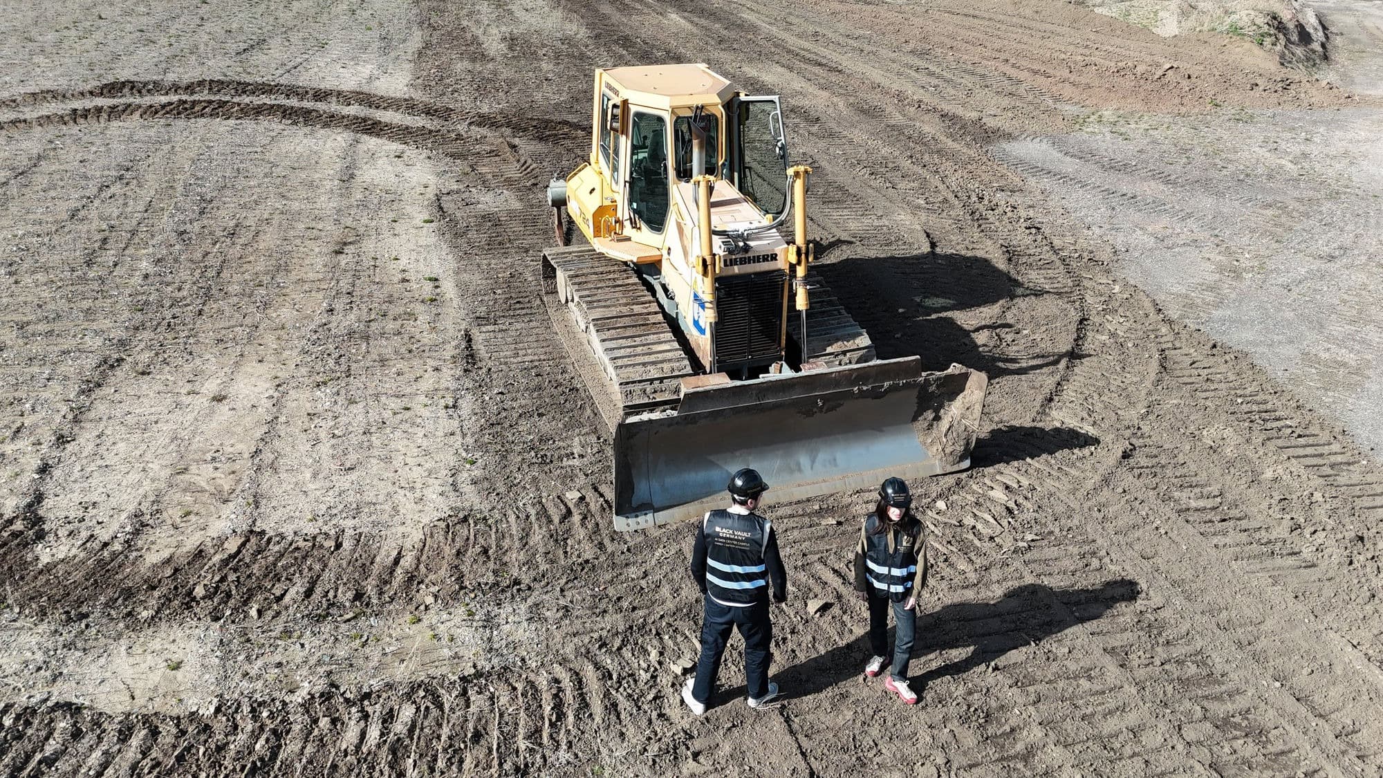 Two people in Black Vault Germany site vests standing before a bulldozer on cleared ground during a site visit.