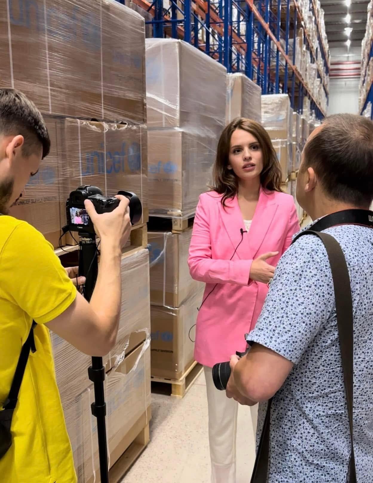Anna Bon in a warehouse of packed medical supplies, speaking to a camera crew.