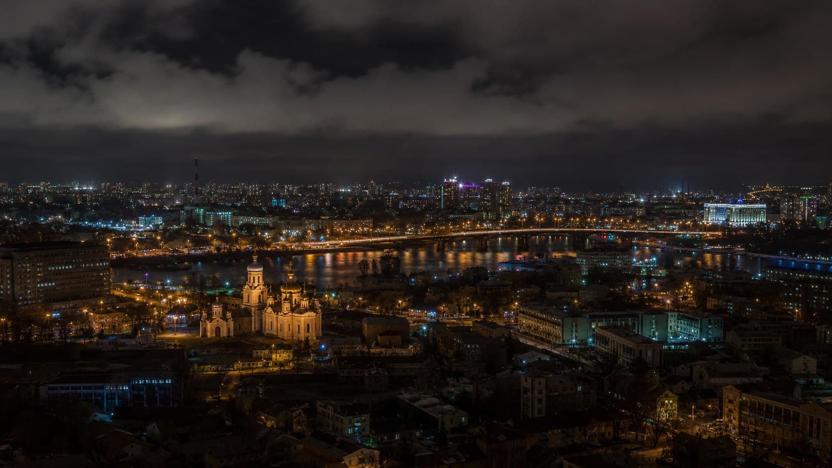 A Ukrainian city at night from above: lit windows, traffic along a bridge, a river reflecting the streetlights.